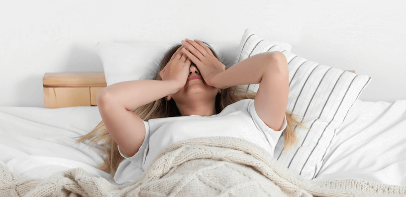 Een vrouw ligt rusteloos in een bed met witte lakens en een beige deken. Haar handen bedekken haar ogen.