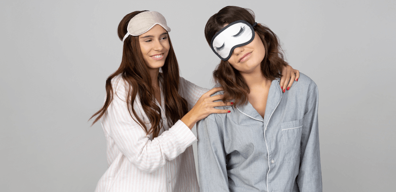 Twee vrouwen in pyjama's en met slaapmaskers. De ene lijkt vrolijk en alert, terwijl de andere er slaperig uitziet en wordt getroost. Tussen hun energieniveau 's ochtends zit een groot contrast.